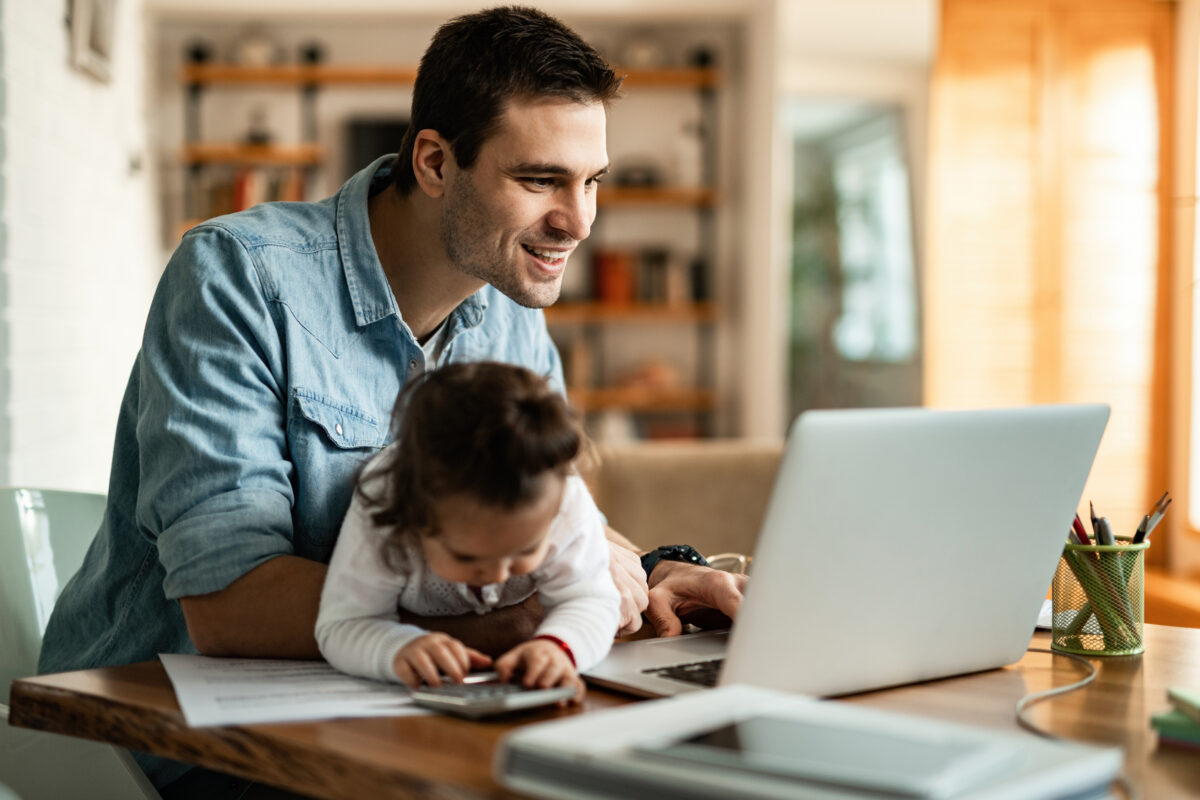 Homem trabalhando home office com a presença de sua filha na mesa de trabalho.