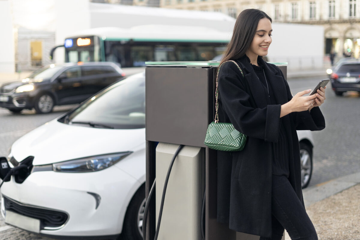 muher mexendo no celular ao lado de seu carro elétrico, que está sendo carregado